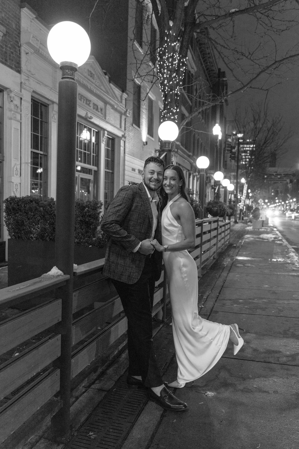 A couple poses on a romantic evening sidewalk with glowing street lamps and twinkling lights in downtown.