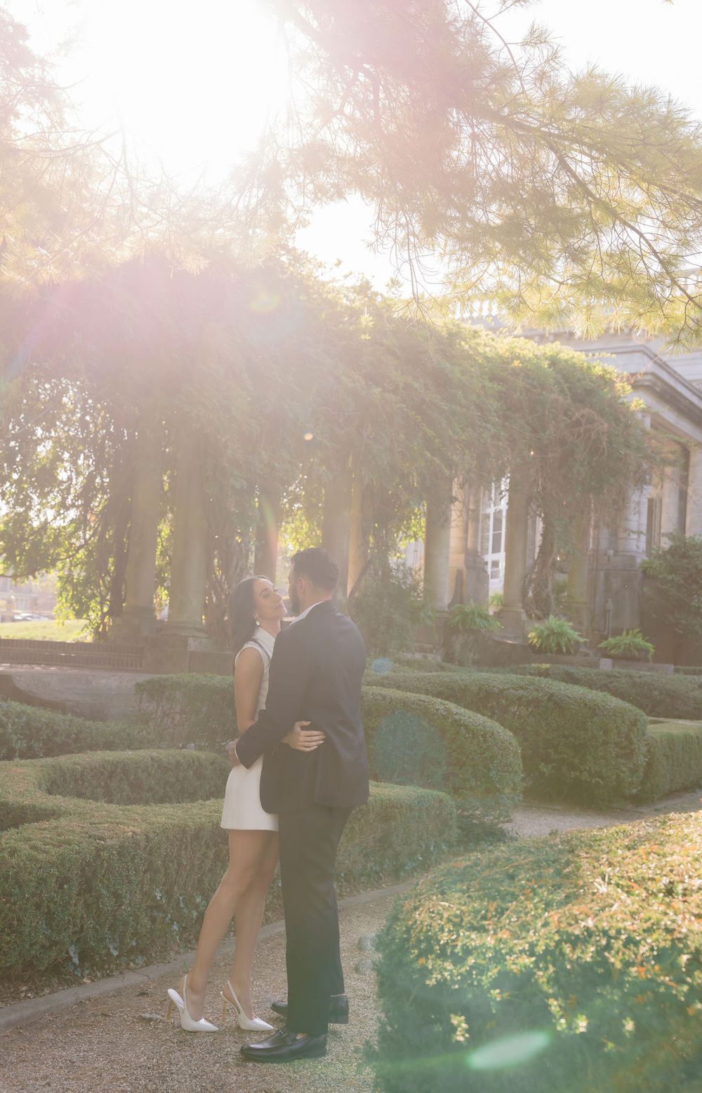 A romantic couple embraces in a sunlit garden with manicured hedges and glowing golden light.