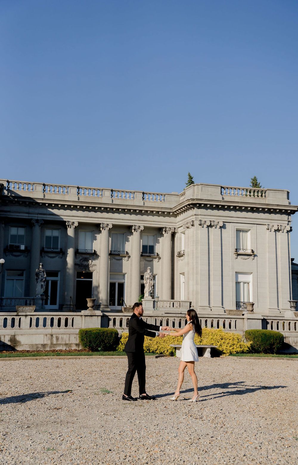 A couple stands in front of a grand neoclassical mansion with white columns and balustrades on a sunny day.