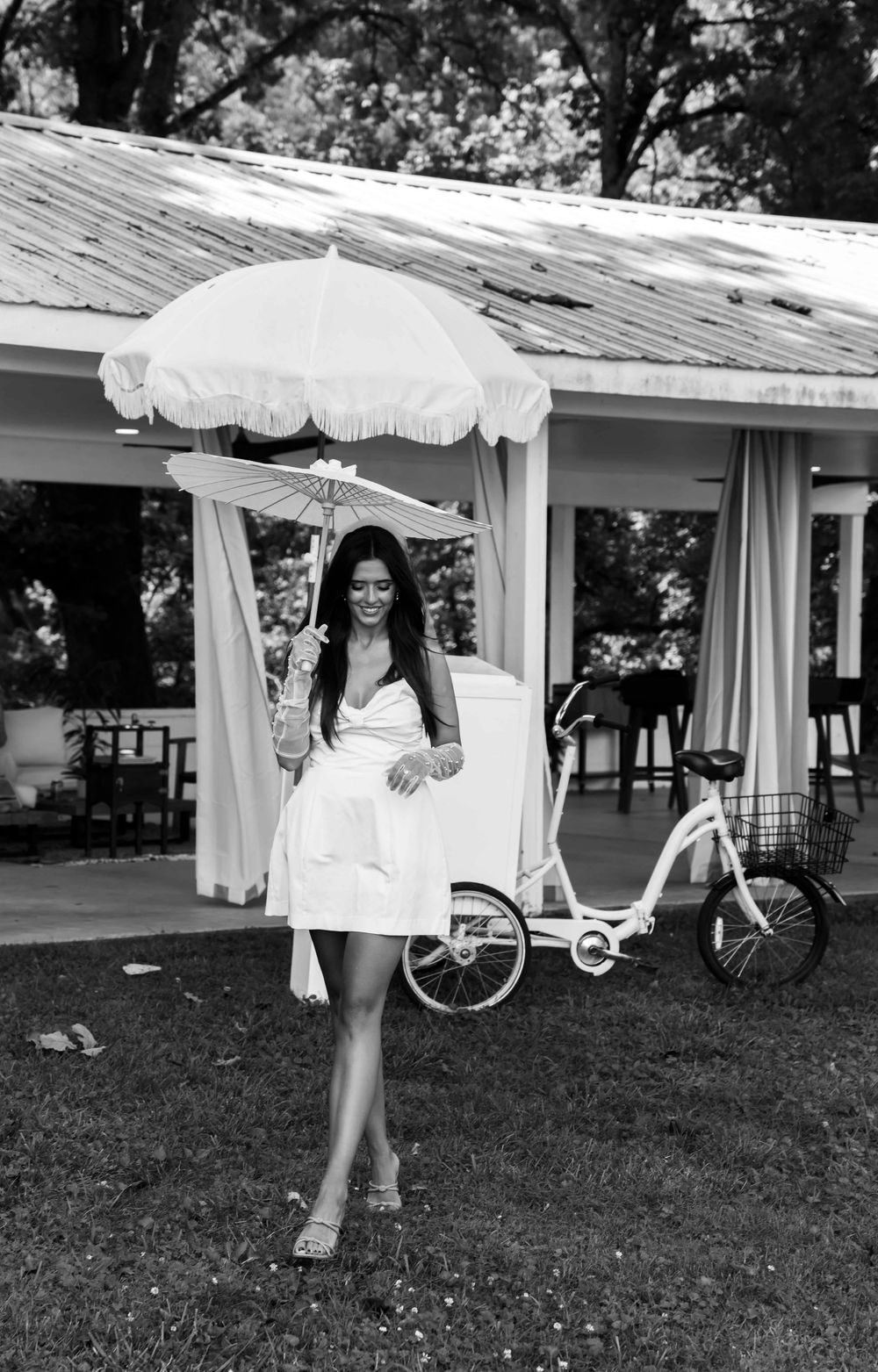 A black and white photo showing a figure in a white dress posing under a white parasol umbrella near a bicycle.