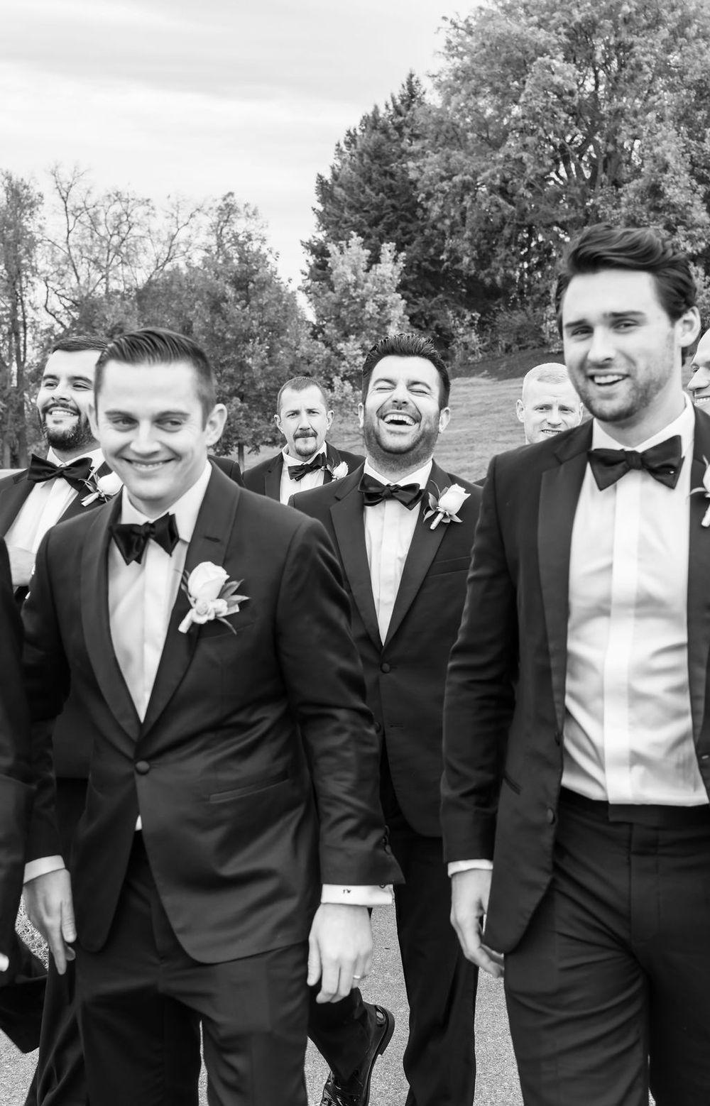 A group of groomsmen in black tuxedos and bowties share a candid moment outdoors.