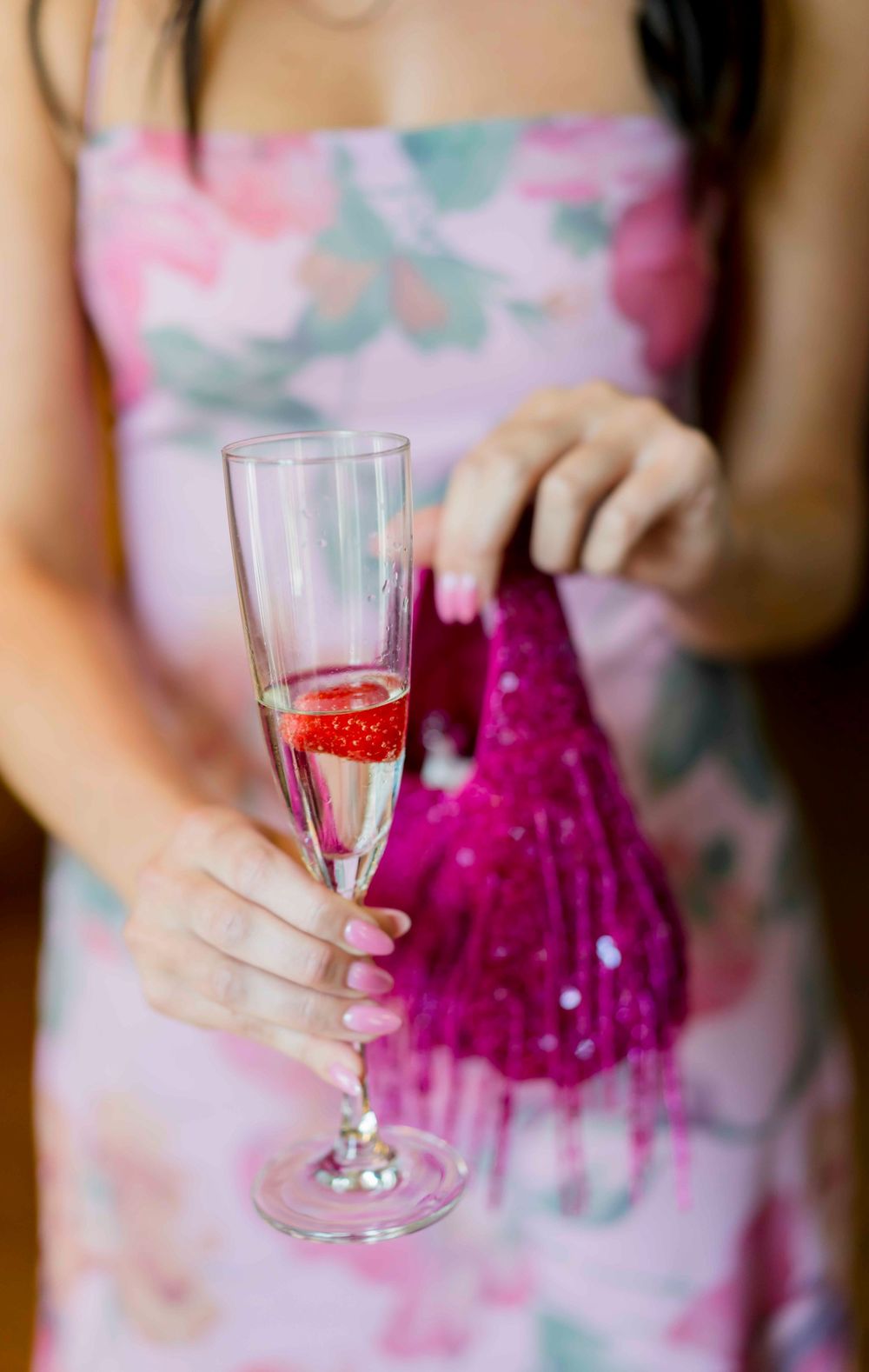 Hands holding a champagne flute with a strawberry garnish next to a bright pink decorative bottle.