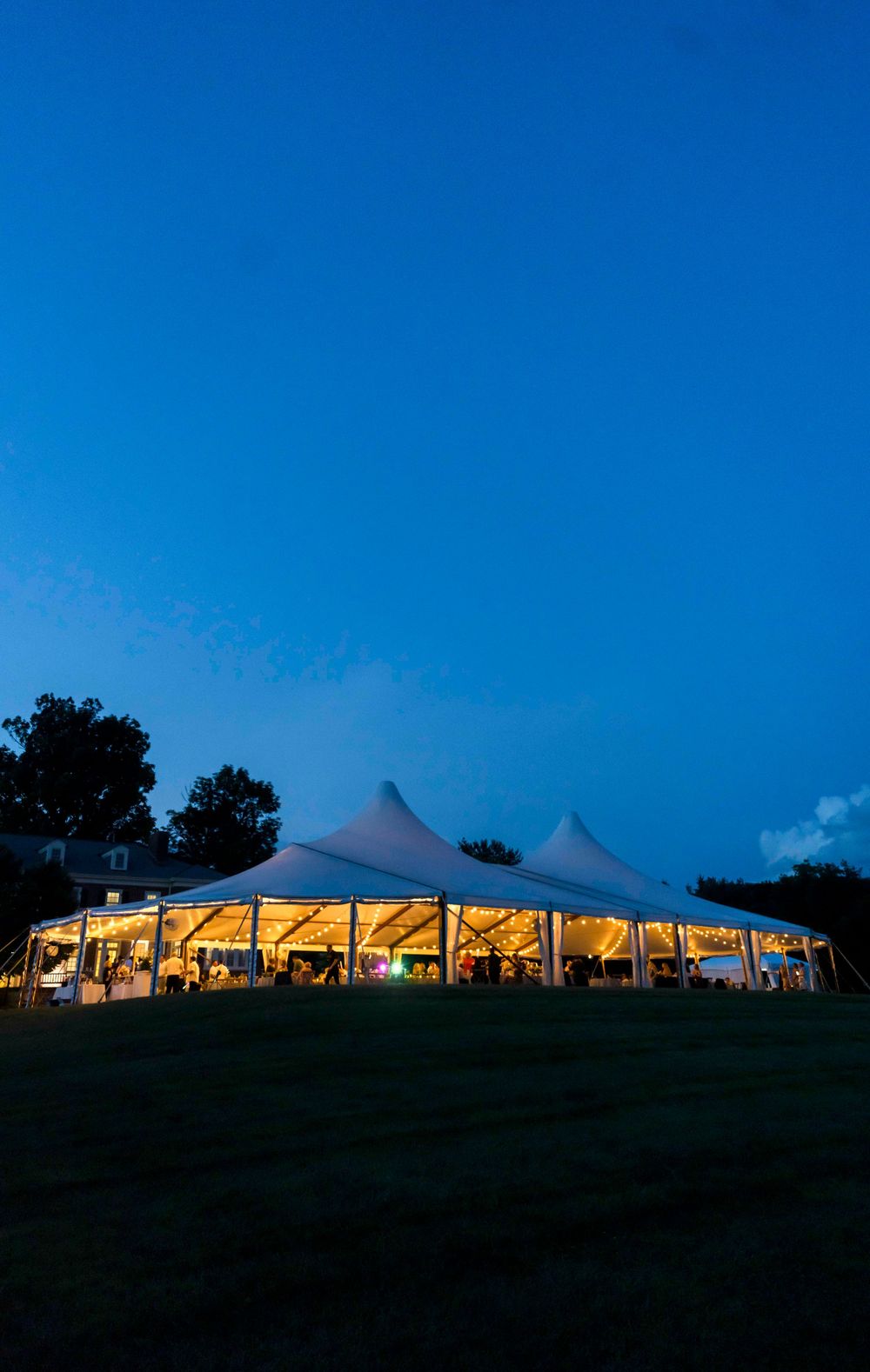 Large white wedding tent glows from within against a dark blue twilight sky at dusk during an outdoor reception.