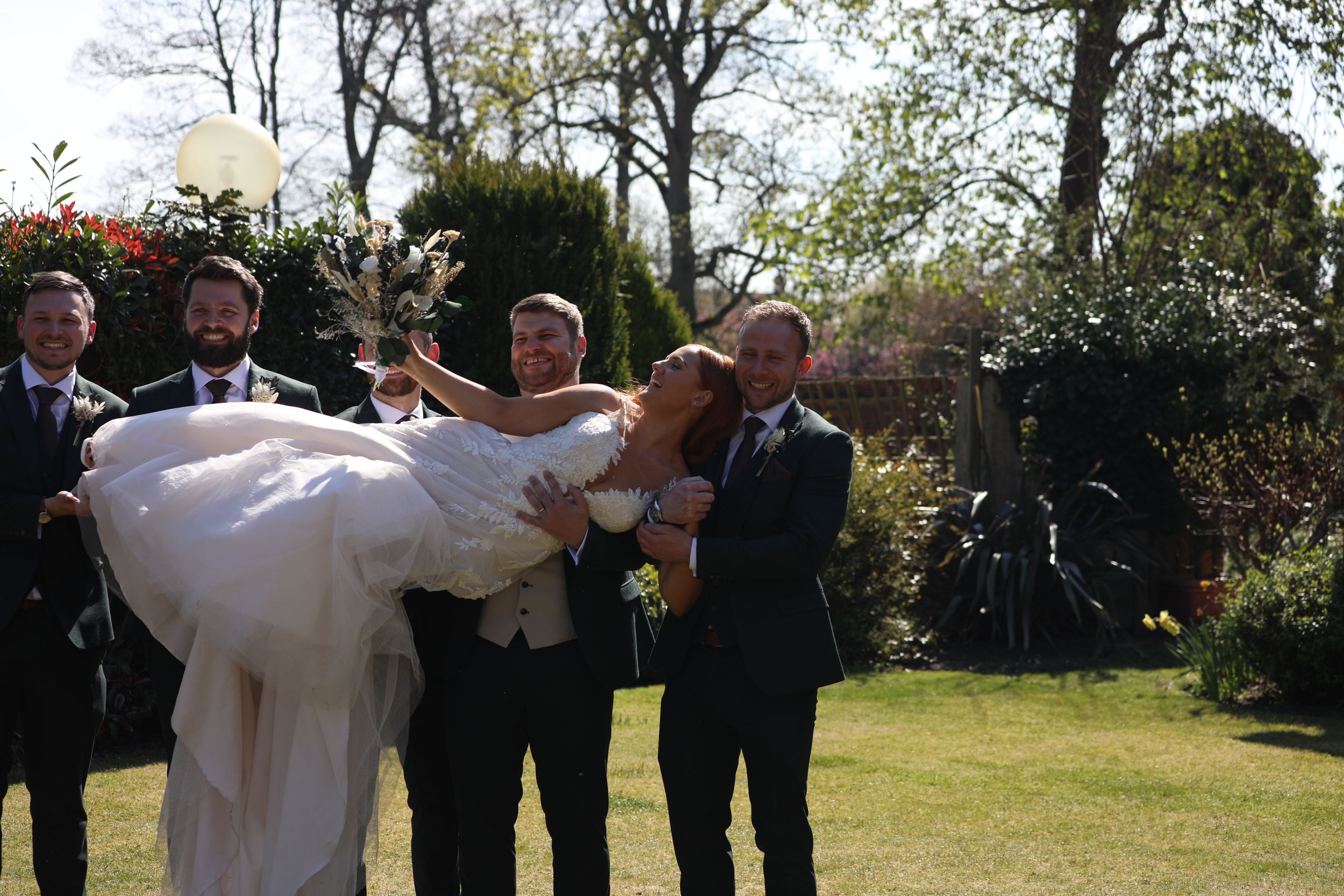Wedding party members lift bride in white dress during outdoor celebration.