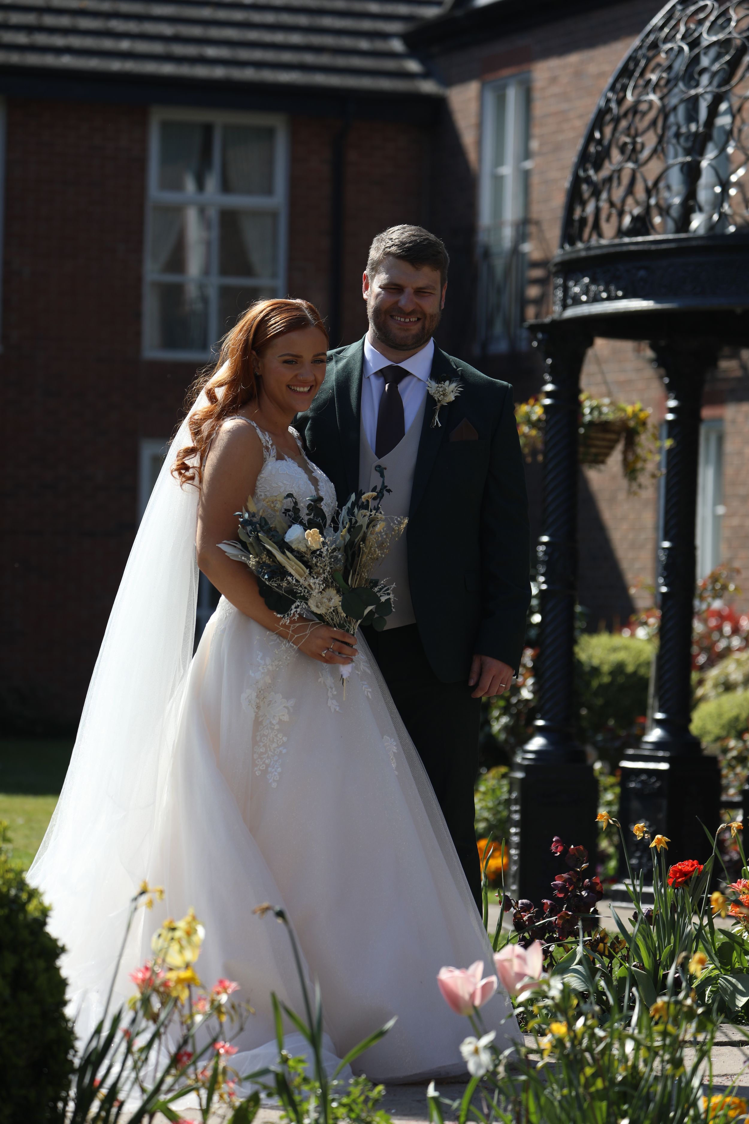 Bride and groom stand together in blooming garden with brick building and decorative gate behind.
