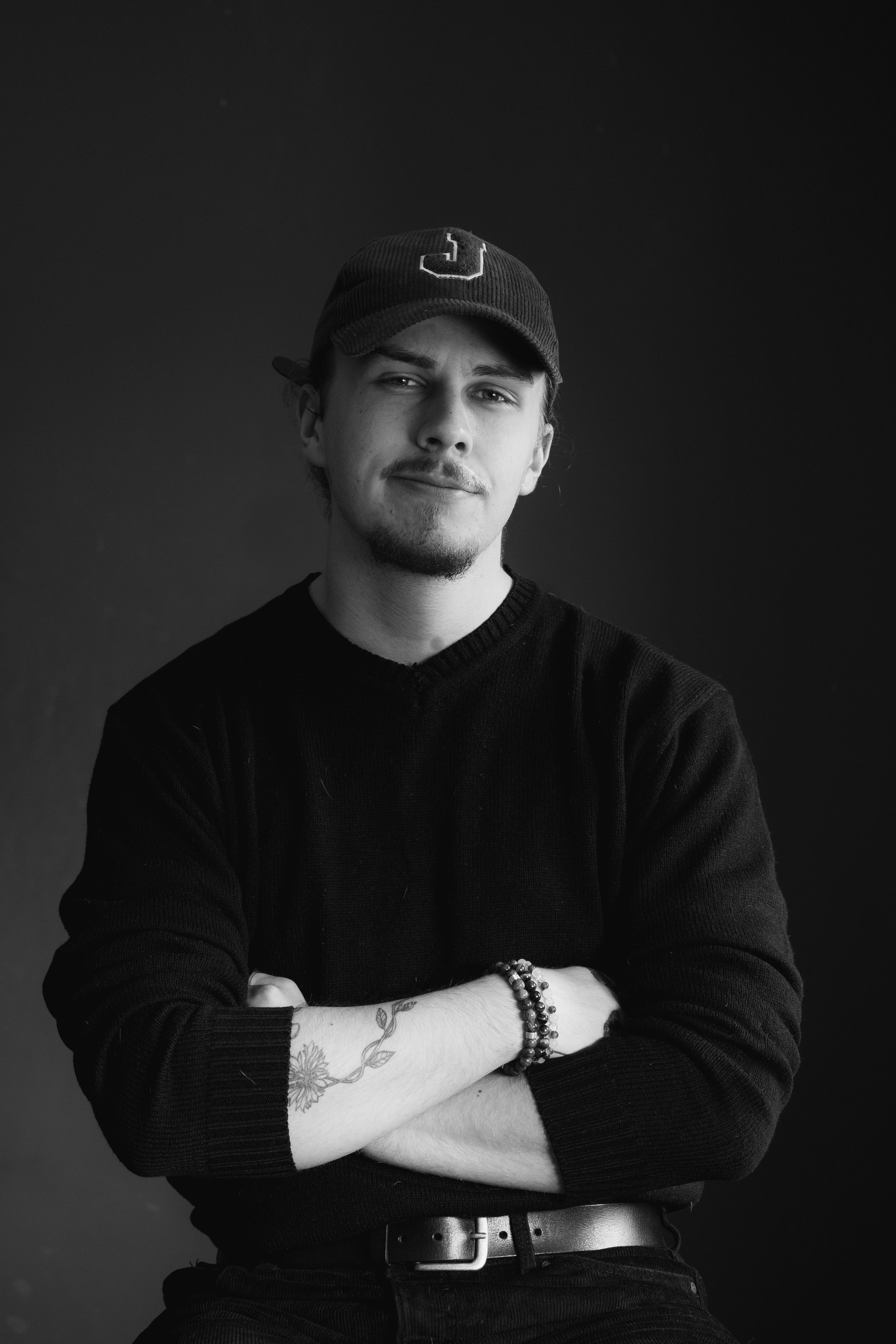Moody black and white portrait of someone in a baseball cap and sweater with crossed arms.