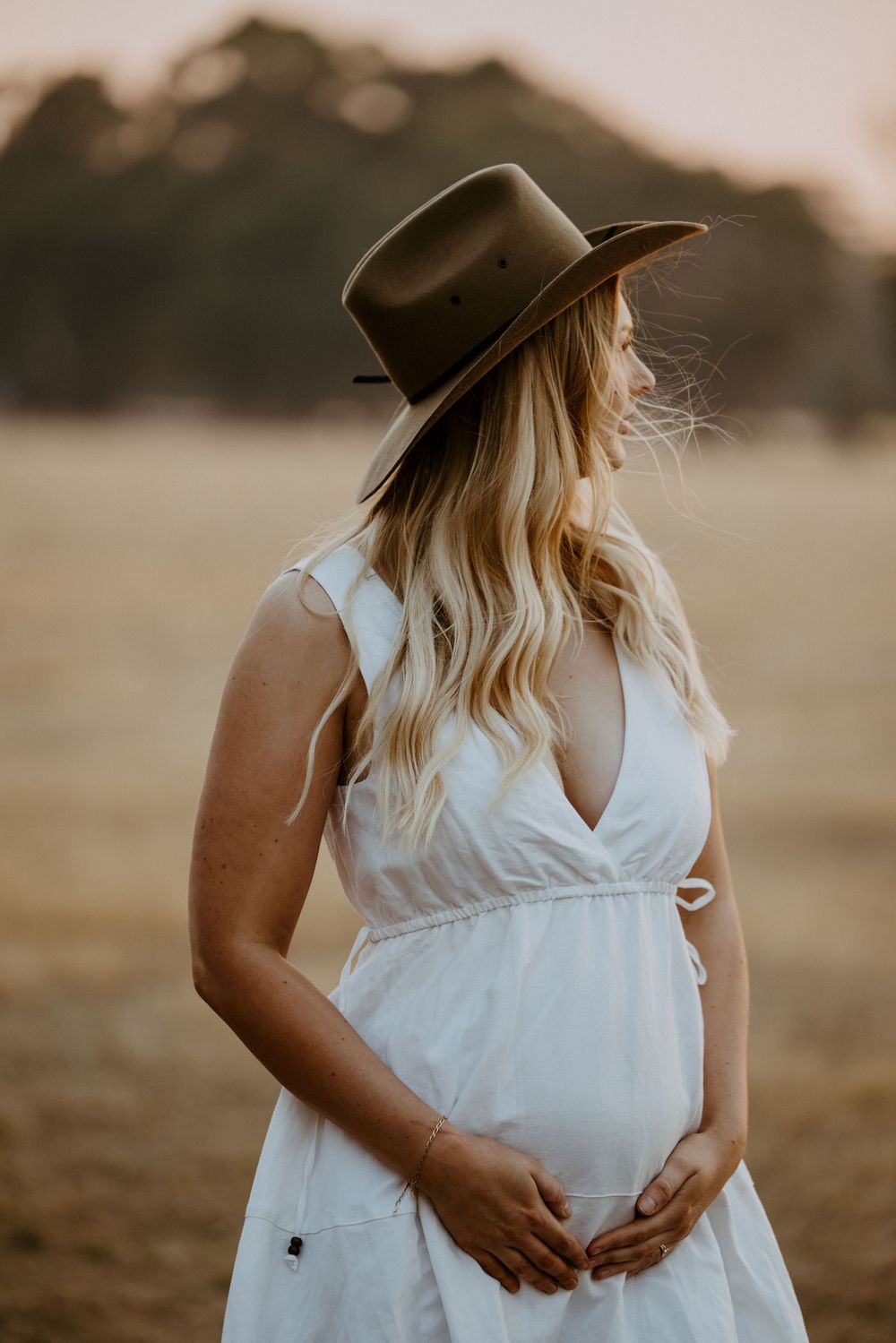 A pregnant woman in a white dress and wide-brimmed hat stands in a golden field at sunset.