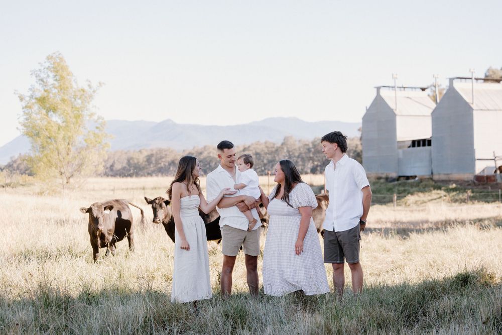 A group in white attire poses together in a rustic outdoor setting with mountains and a white building in the background.