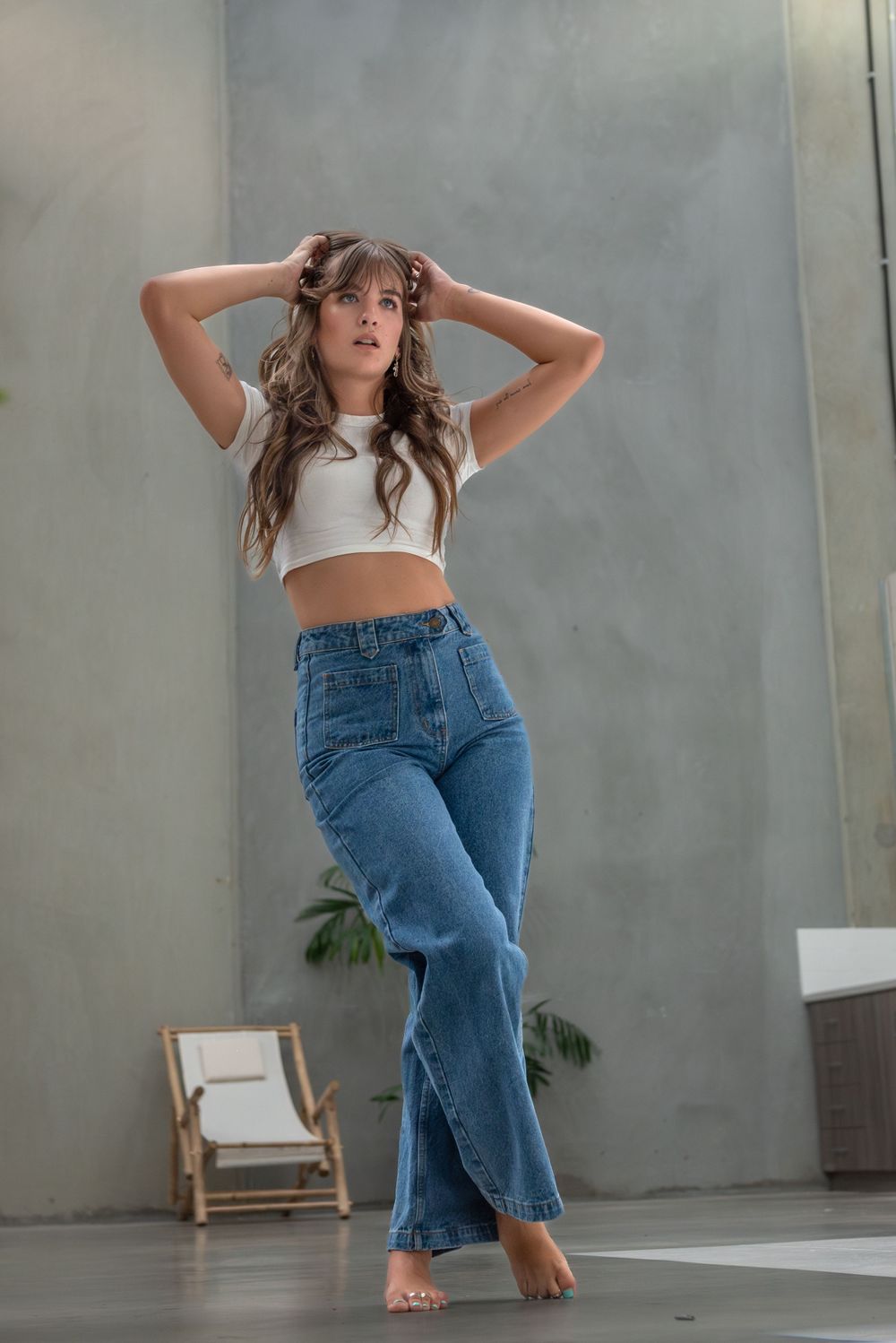 Young woman in crop top and jeans poses casually against grey wall.
