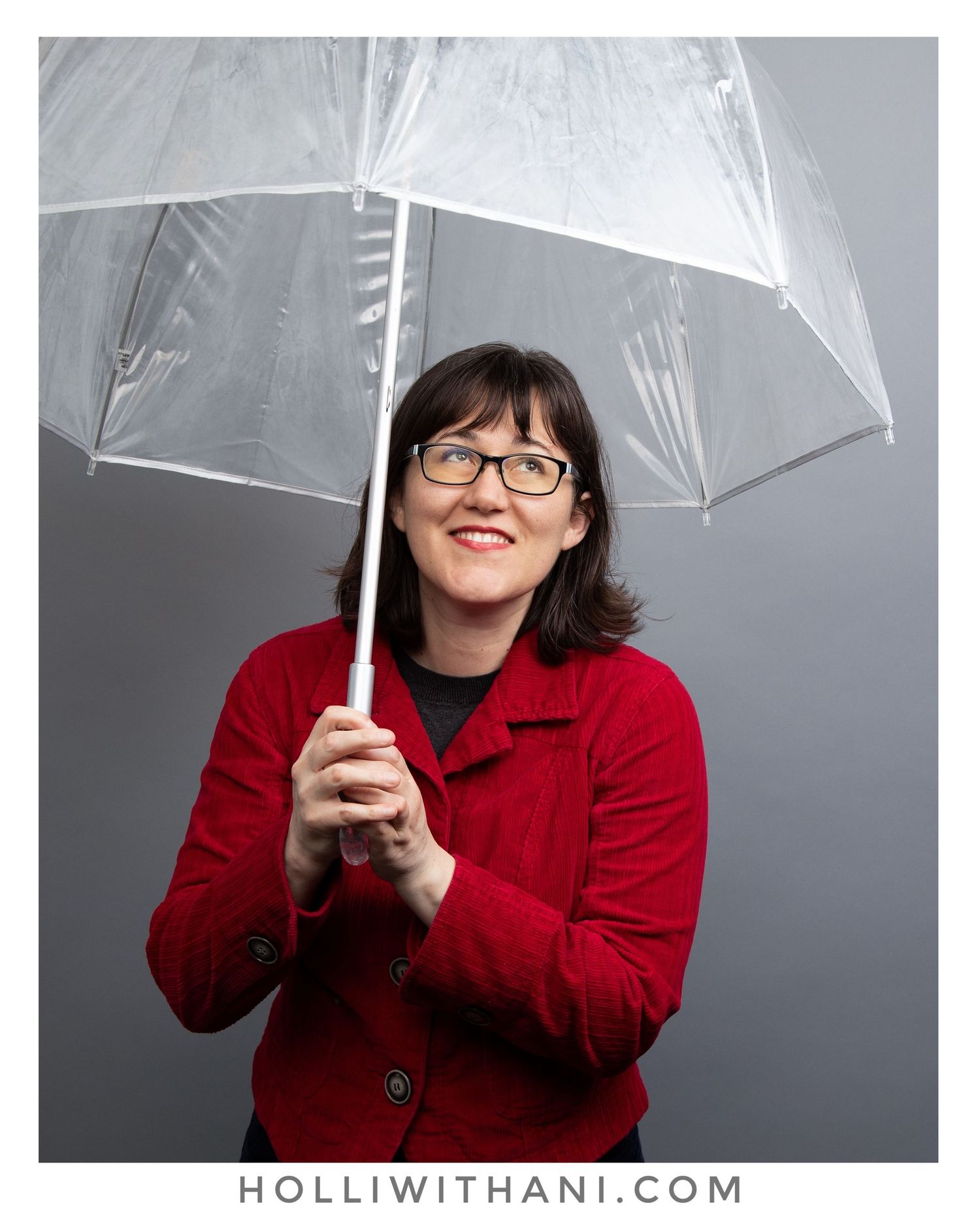 Color photo of woman with clear umbrella and gray backdrop.