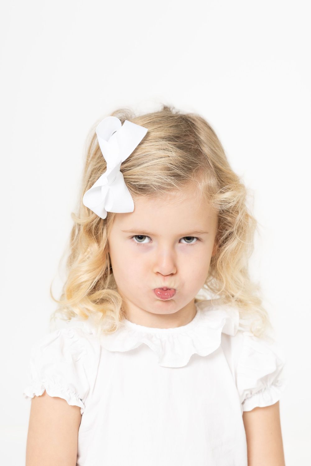 A young child wearing a white dress and bow makes a pouty expression against a white background.