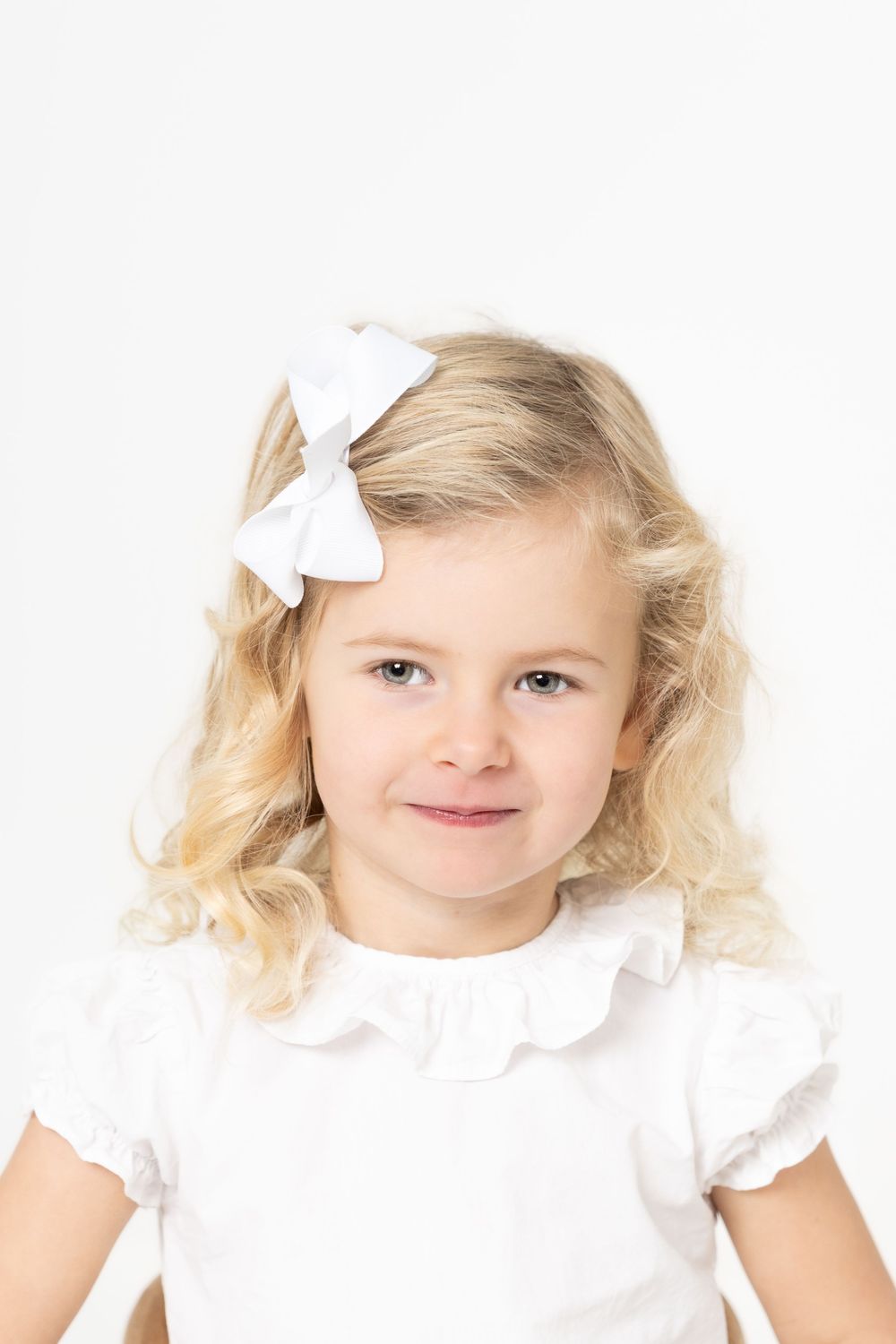 Portrait of a young child with blonde curly hair wearing a white top and white bow hair accessory against white background.