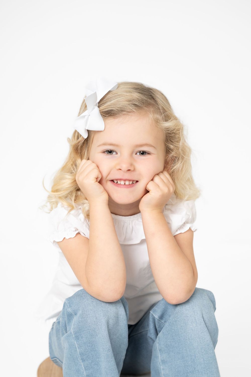 Child in white top and jeans sits with hands propped under chin in playful pose against white background.