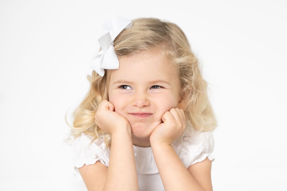 Child with blonde curls wearing white dress and hair bow poses with hands on cheeks against white background.