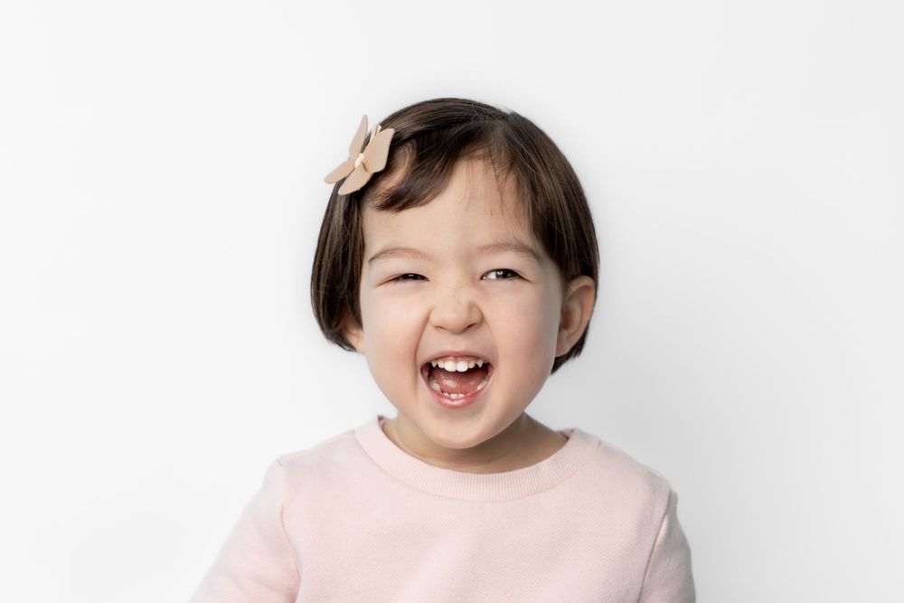 A joyful young child in a pink shirt laughs and smiles while looking at the camera against a white background.