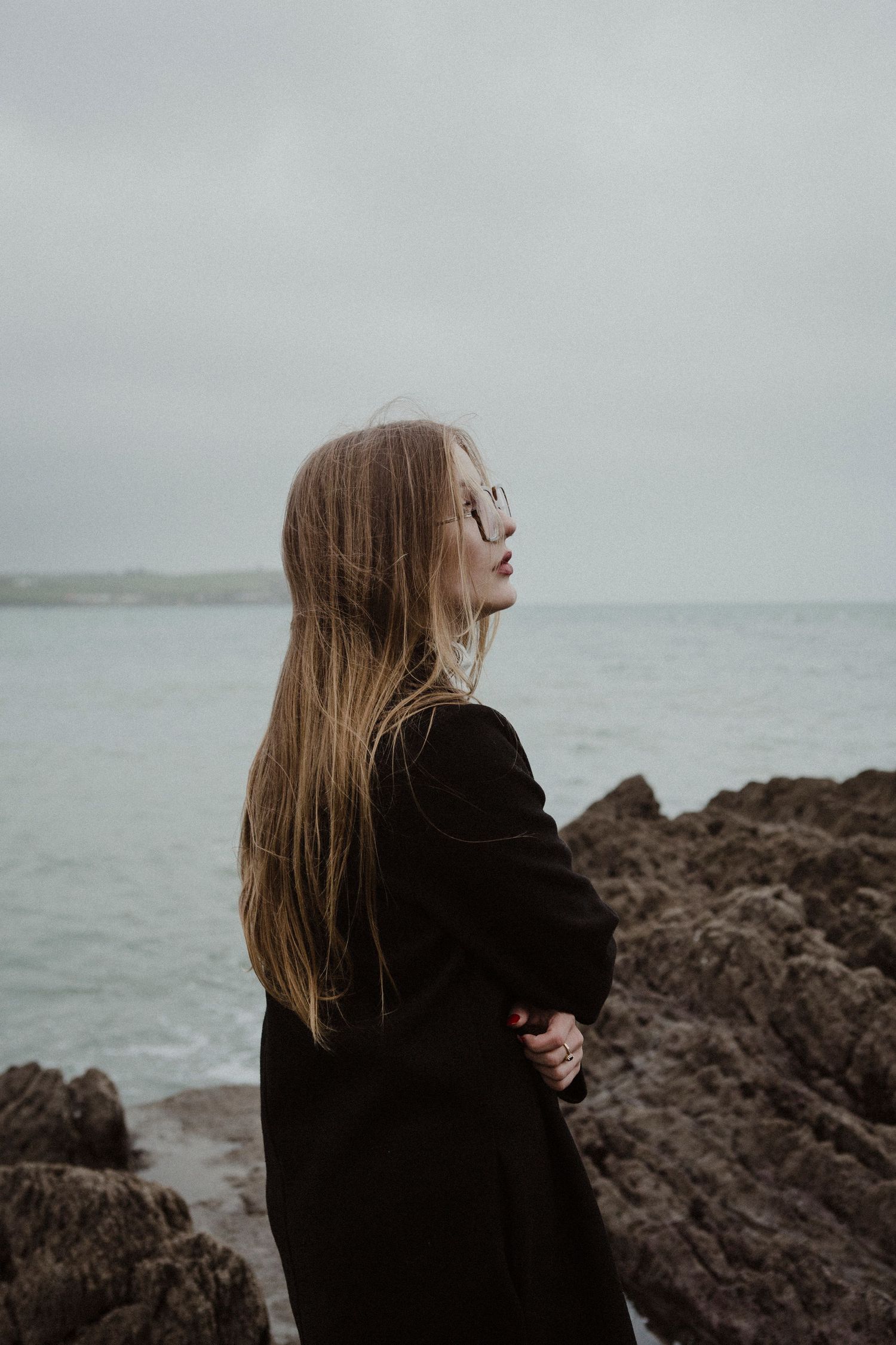 Figure with long hair poses thoughtfully against rocky coastal backdrop in dark winter coat.