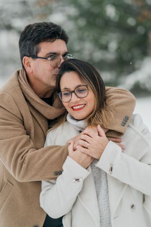 A couple wearing glasses and winter coats embrace in a tender moment against a snowy outdoor backdrop.