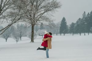 A person in a red winter hat walks through a snowy park landscape carrying shopping bags in winter weather.