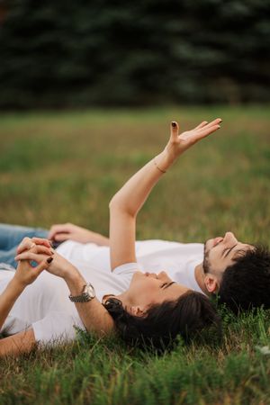 A couple lying in grass with raised arms reaches towards the sky during golden hour.