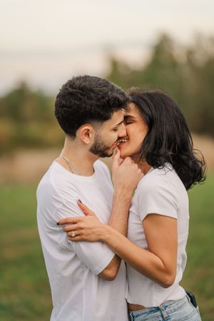 Romantic couple in white shirts share tender kisses while embracing in a grassy field during sunset.