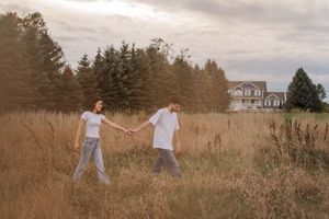 Couple holding hands and walking through tall grass with evergreen trees in background.