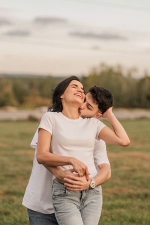 A couple in white t-shirts and jeans share playful embraces and laughter in a sunny outdoor field.