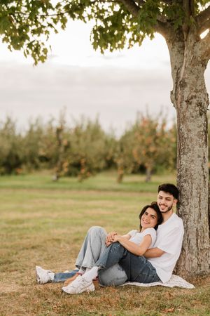 A couple relaxes together under a shady tree in an orchard on a sunny afternoon.
