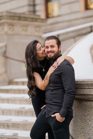 A romantic couple in black outfits share tender moments on stone steps during an engagement photoshoot.