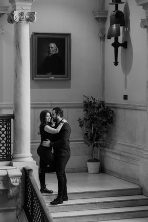 A romantic couple embraces on a grand staircase in a classic building interior with white columns and crosses on display.