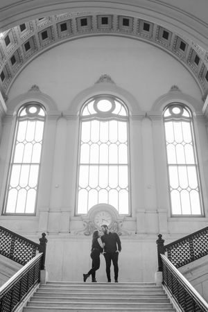Two silhouettes ascend a grand marble staircase in a historic building with arched windows and ornate architectural details.