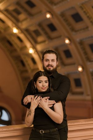 A couple in formal black attire poses together on a curved architectural structure with warm lighting.
