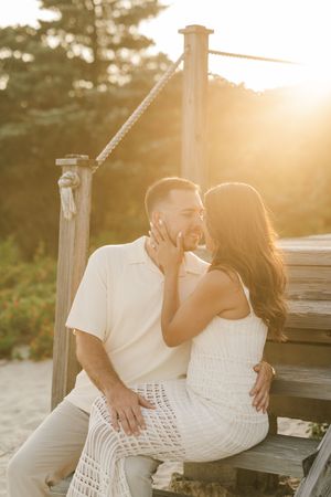A romantic sunset beach photoshoot with a couple in white outfits sitting on a wooden bench.