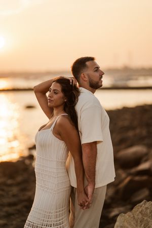Romantic couple in white outfits posing against rocky beach landscape during sunset.