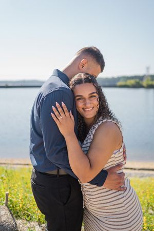 A couple embraces on a sunny day by a waterfront with rolling hills in the background.