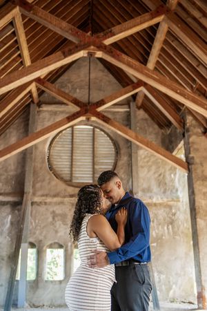 A couple shares an intimate moment under rustic wooden beams in an old building with weathered walls.