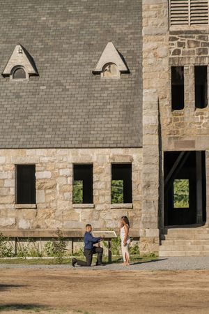 A romantic proposal moment takes place outside a historic stone building with dormer windows on a sunny day.