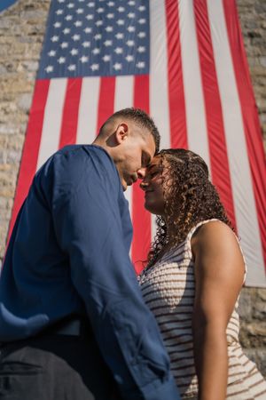 A romantic couple shares a tender moment in front of American flags against a stone wall backdrop.