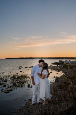 Couple in white clothing stands by rocky shoreline during sunset over calm waters.