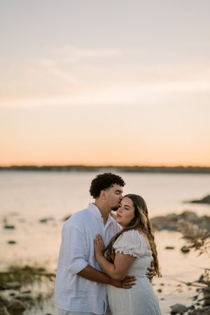 A romantic couple embraces during sunset at a rocky coastal beach with warm orange sky in the background.