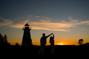 Silhouetted figures dance in front of a lighthouse against a vibrant orange sunset sky.