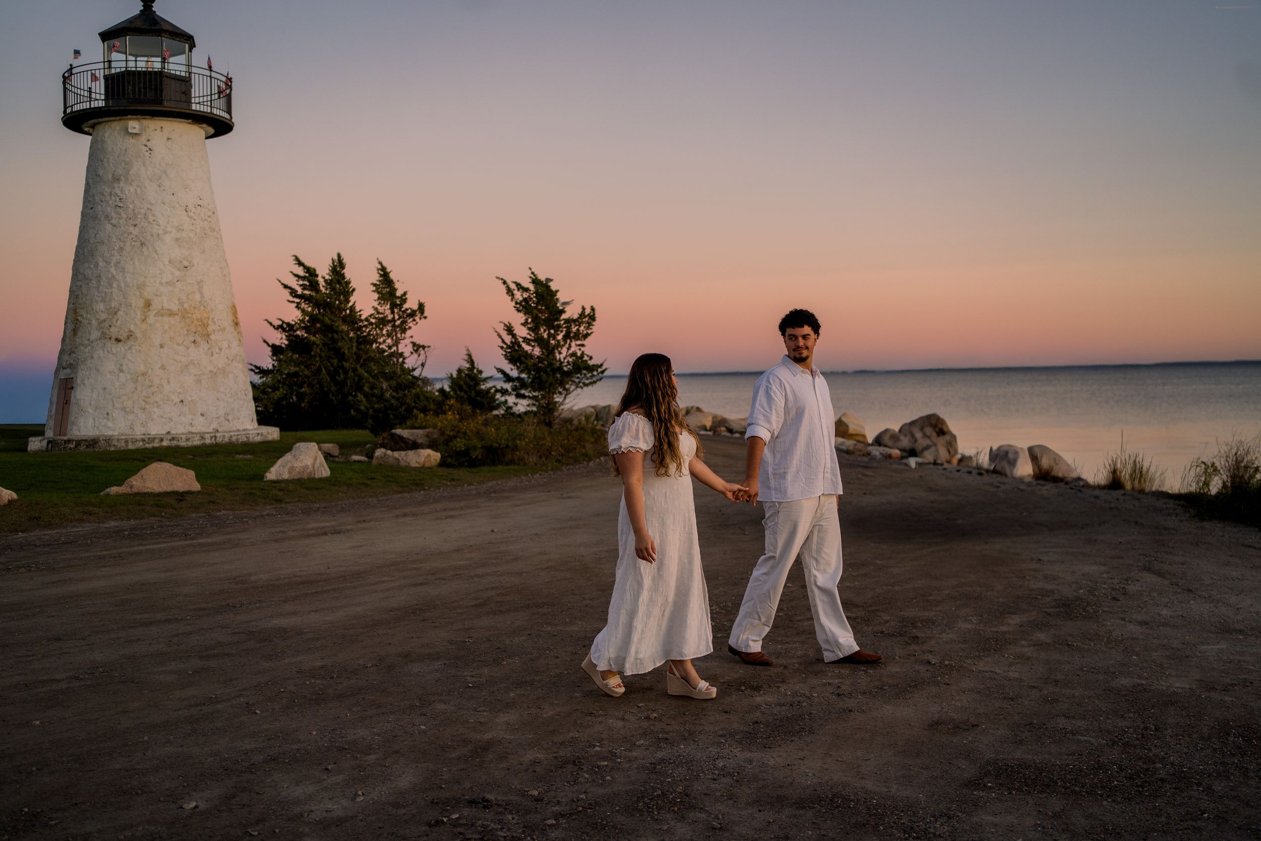 A romantic lighthouse engagement photo session at sunset with a couple in white outfits by the ocean.
