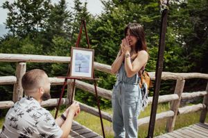A surprise proposal moment captured on an outdoor wooden deck surrounded by trees.