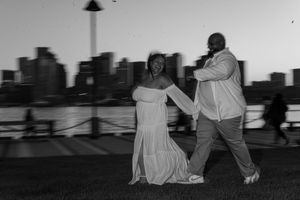 A couple dances together on a lawn with a city skyline silhouetted in the background at dusk.
