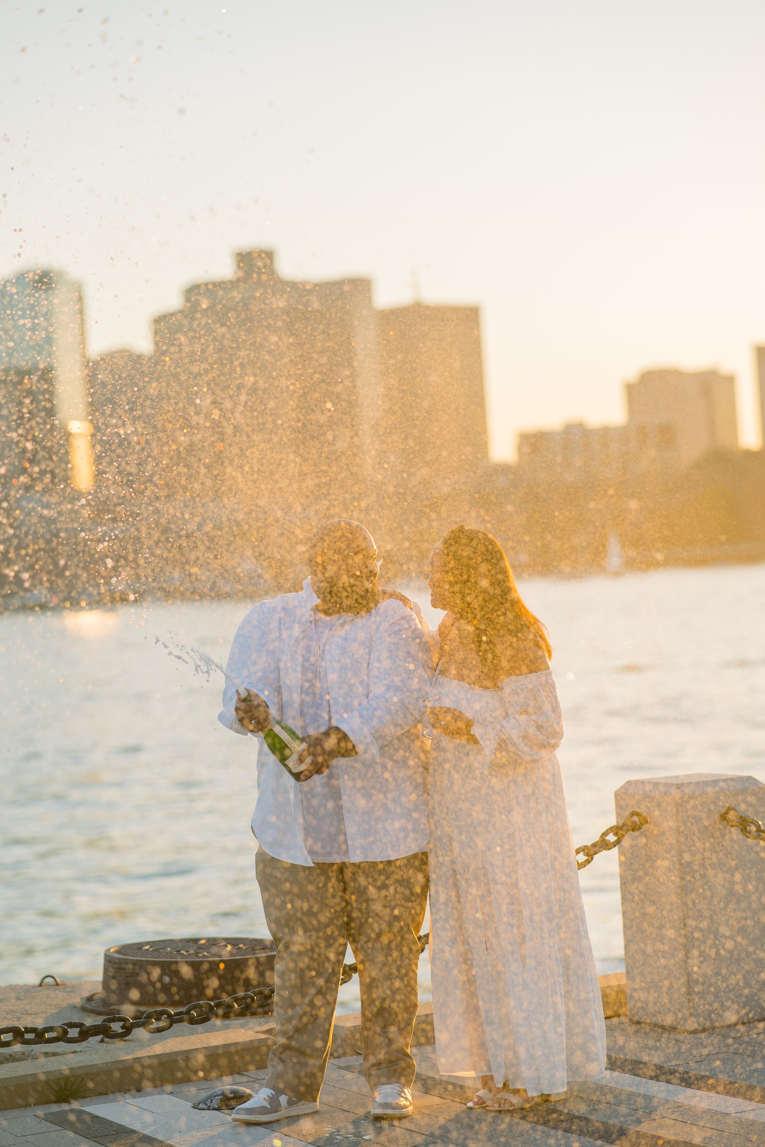 A couple embraces while watching the sunset over a city skyline from a waterfront location.