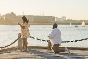 A couple sits together on a wooden dock at sunset overlooking a harbor with boats in the distance.
