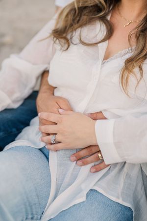 Close-up view of hands wearing engagement ring resting on white fabric in soft natural light.
