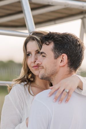A romantic couple in white shirts embrace while sharing an intimate moment under an outdoor structure.