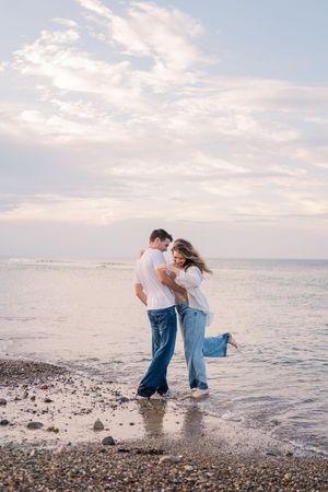 A couple playfully dips and poses along a rocky beach shoreline at sunset with ocean waves in the background.