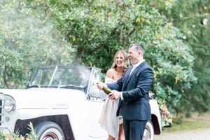 A couple poses beside a vintage white car surrounded by greenery.