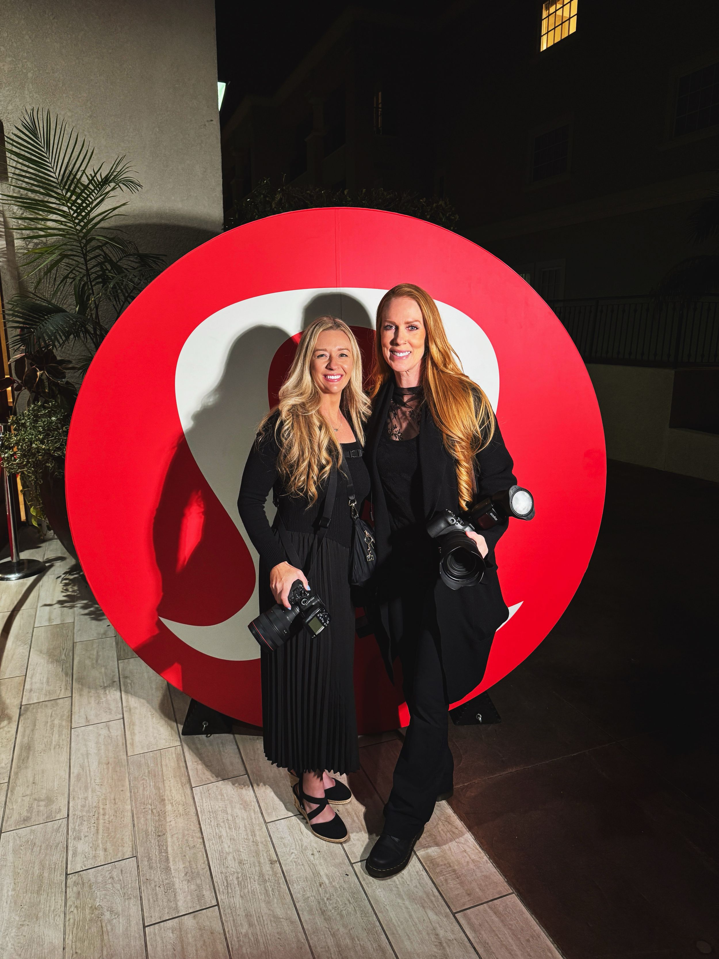 Photographers posing together against large red decorative backdrop in stylish black outfits.